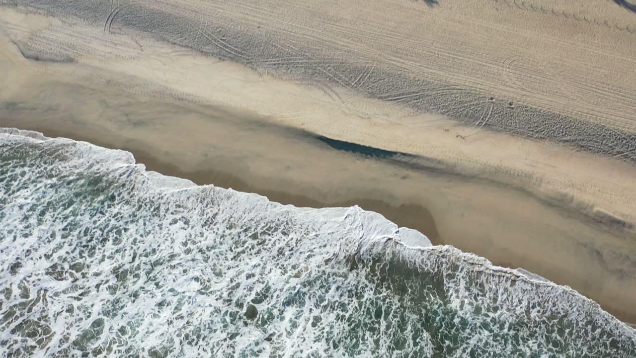 Aerial Los Cabos beach frame with foamy water at the bottom, darker wet sand near the shore, and wide pale sand stretching toward the top, with no visible people.