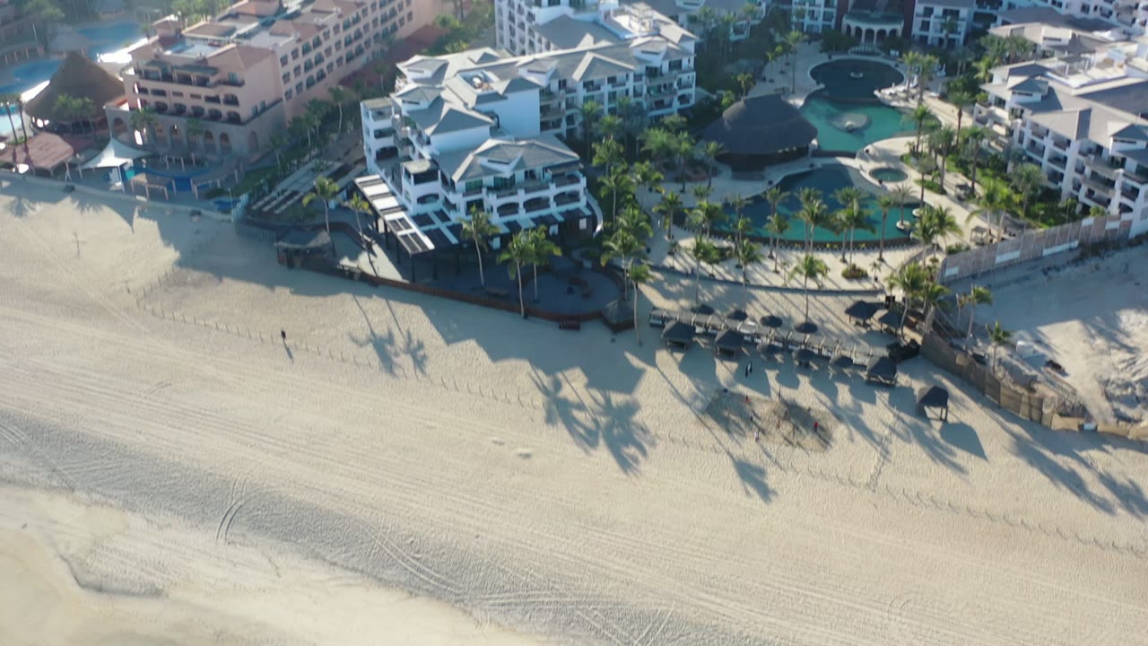 Opening aerial frame over Los Cabos, Mexico beachfront hotels with turquoise pools, palm shadows across the sand, and no discernible people visible.