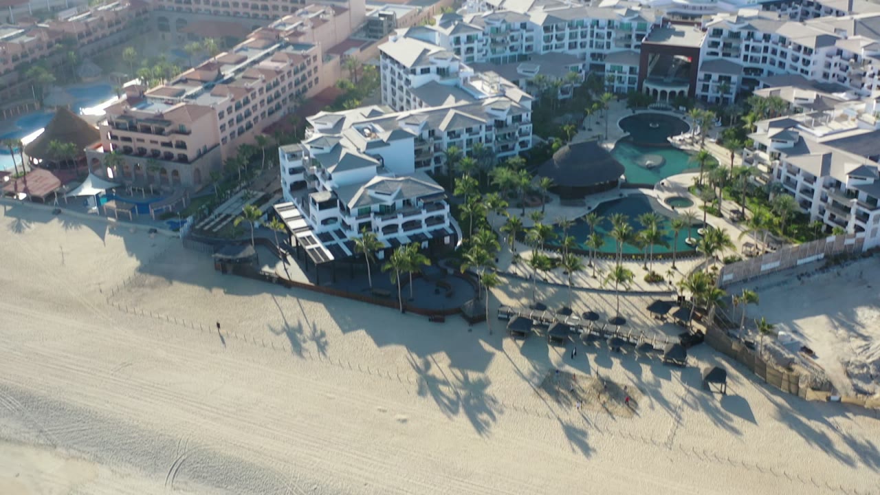 Aerial frame in Los Cabos, Mexico showing white and peach hotel buildings beside pool courtyards, beach cabanas, and sunlit sand with no discernible people visible.
