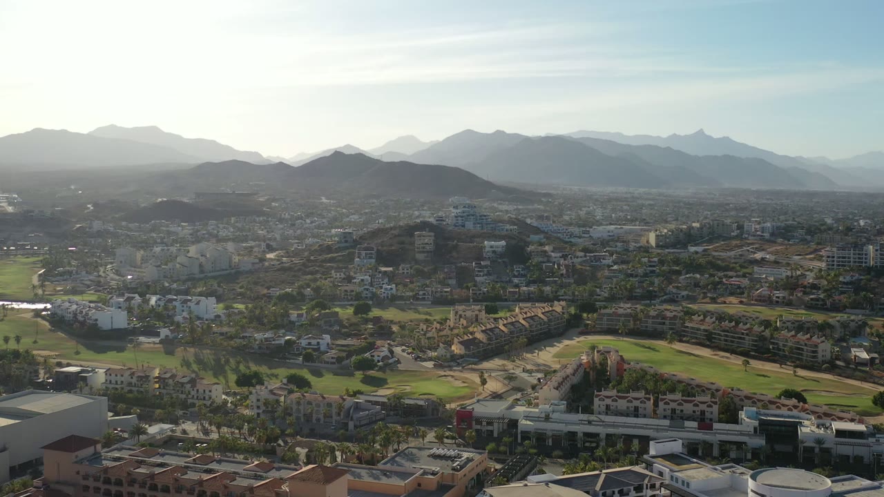 Second frame of the Los Cabos aerial view, showing a wide urban area with green fairway-like strips, clustered buildings, a hazy mountain range, and no visible people.