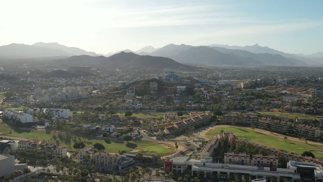 Wide aerial frame over Los Cabos, Mexico, with terracotta-roof homes, green landscaped areas, roads, and hazy mountain ridges in the distance, with no visible people.