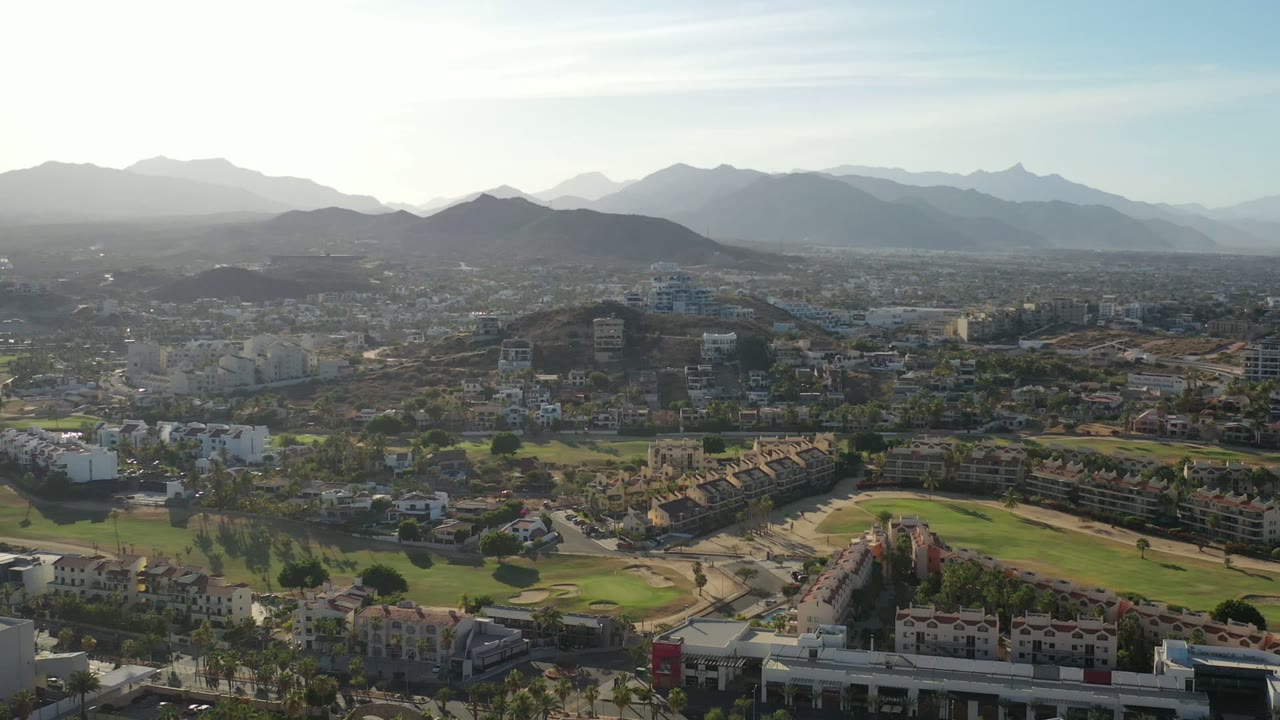 High aerial view in Los Cabos, Mexico, showing white buildings, curving green fairways, roads, palm-lined areas, and layered mountain silhouettes, with no visible people.
