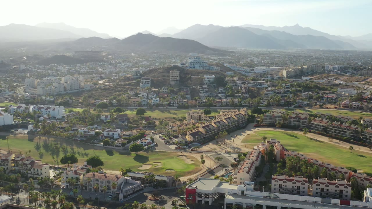Opening aerial frame over Los Cabos, Mexico, with red-roofed homes, green fairways, clustered buildings, hazy mountains, and no visible people.
