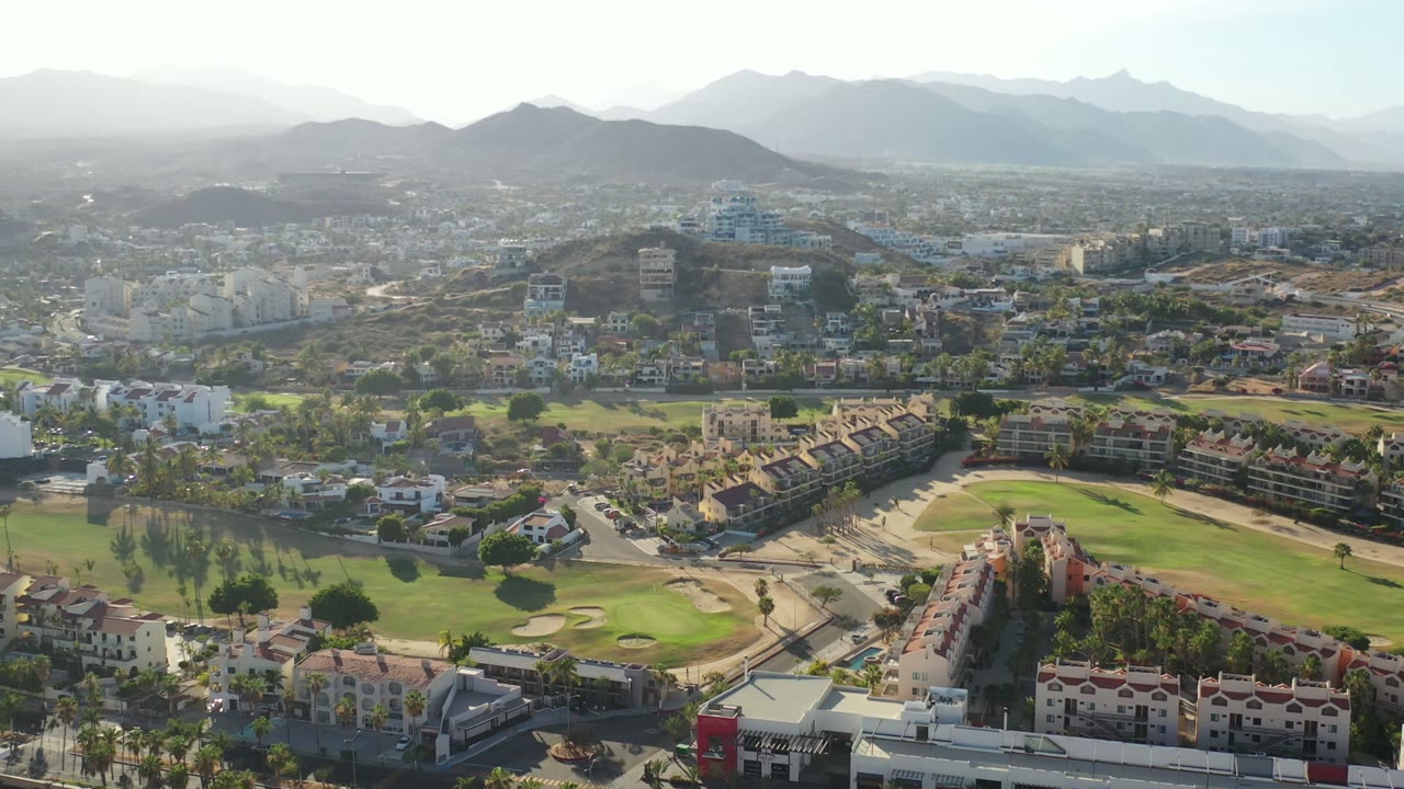 Second Los Cabos aerial frame looking over residential rows, a central green fairway, pale city buildings, distant hills, and no visible people.
