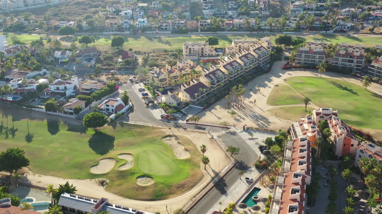 Second aerial frame in Los Cabos showing a brighter fairway and bunkers in the lower view, intersecting roads, rows of homes, background hillside development, and no visible people.