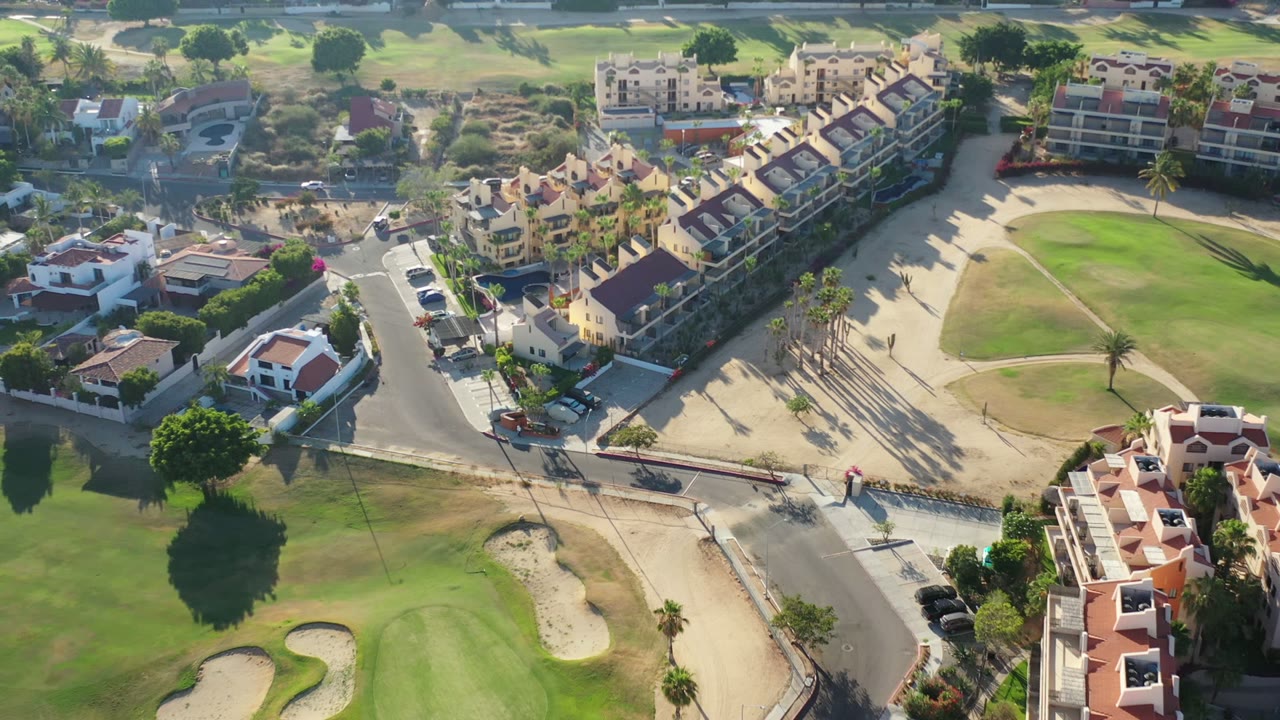 Oblique drone view over Los Cabos, Mexico, with red-roofed homes, a curved road, fairways, sand traps, palm trees, and no visible people.