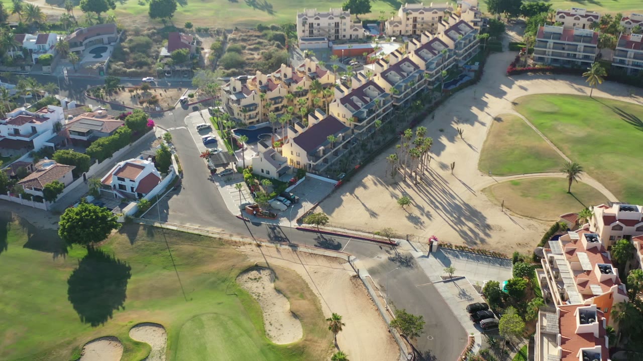 Aerial frame in Los Cabos, Mexico, showing a road junction between residential buildings and a bright green fairway, with no visible people.