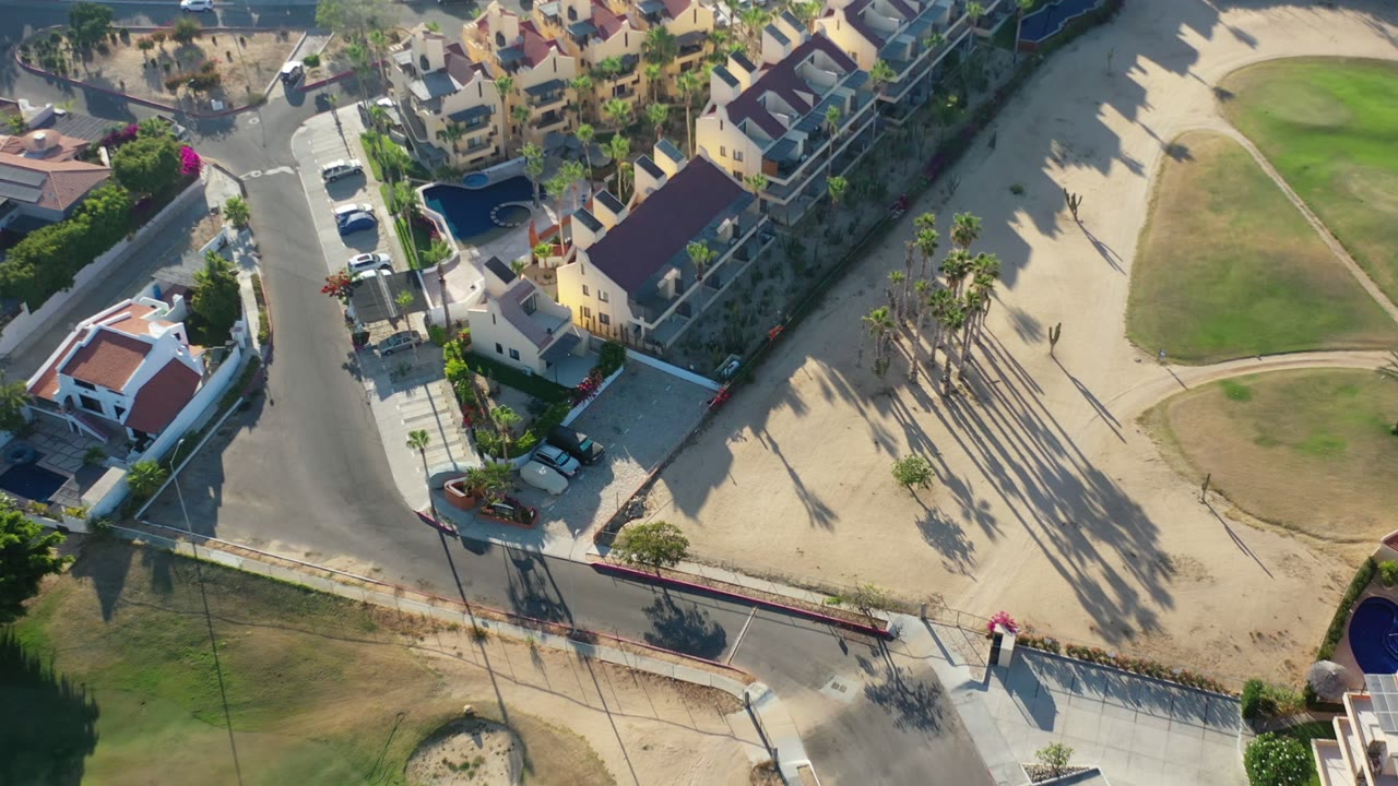 Overhead frame from Los Cabos showing a curved gray street, walled homes, tan sand, a green lawn edge, tall palm shadows, parked cars, and no visible people.