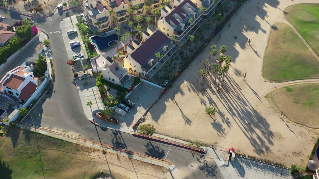 Los Cabos aerial frame with clustered homes along gray roads, parked vehicles near driveways, sandy terrain, a rounded green lawn section, palm trees, and no visible people.