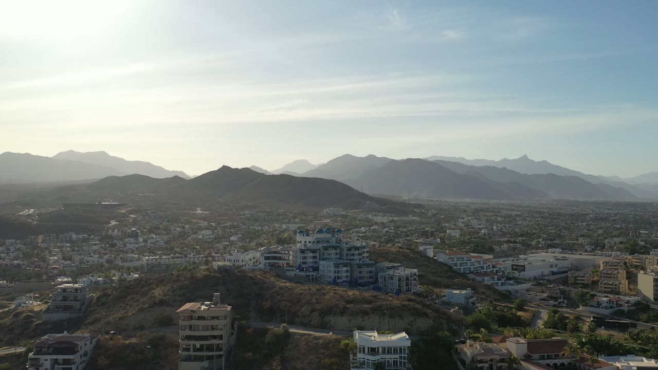 Wide aerial view of hillside buildings, city, and distant mountains under bright sky.