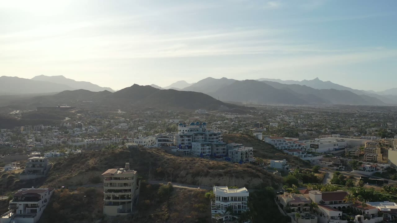 Los Cabos aerial frame with multi-story hillside buildings near the lower center, city blocks stretching across the valley, pale mountains layered behind them, and no visible people.