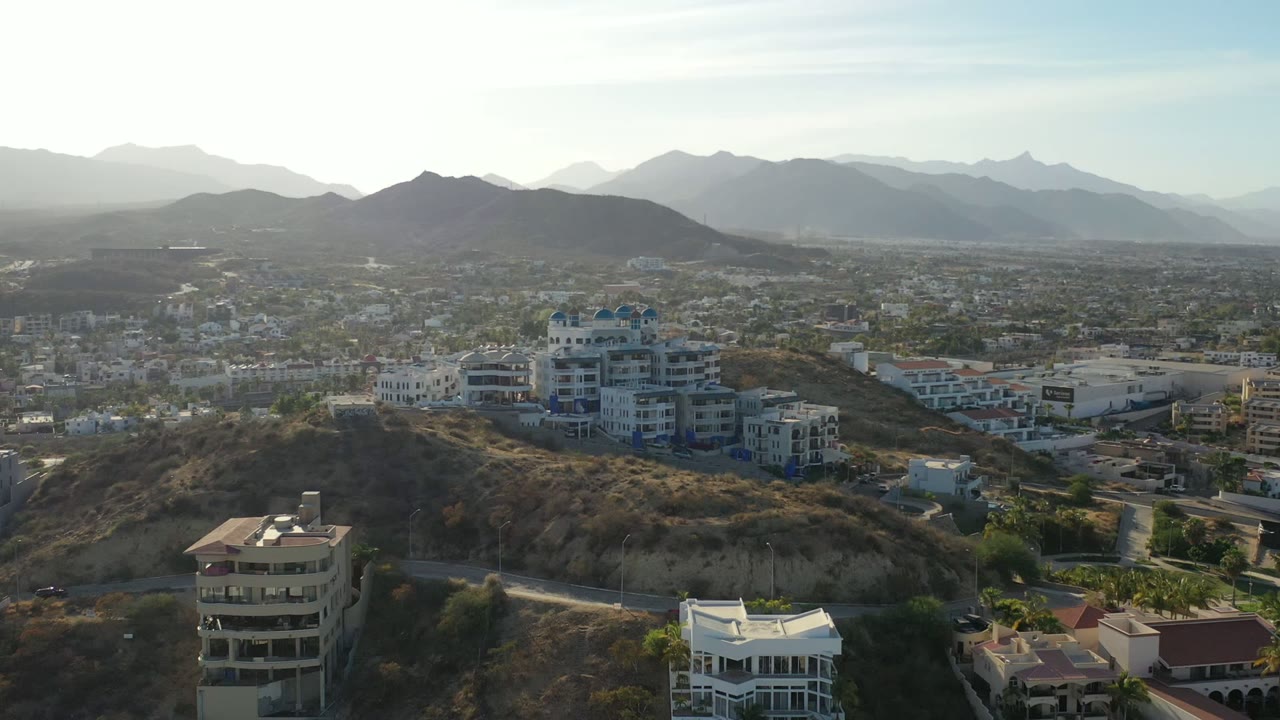 Second aerial frame over Los Cabos, Mexico showing a round foreground building, white villa rooftops, a dry hill, city blocks, distant ridges, and no visible people.