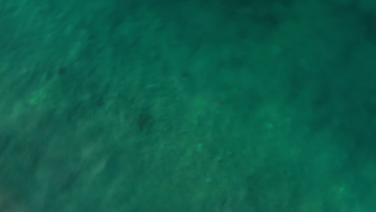 Overhead Los Cabos ocean frame showing green-blue water, slight ripple streaks, scattered darker patches below the surface, and no visible people.