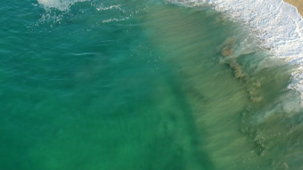 Overhead Los Cabos water frame with a brighter white wave curling into the upper right and green shallows visible below, with no visible people.