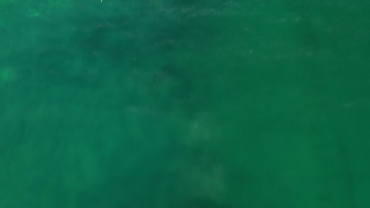 Aerial frame of Los Cabos ocean water with a broad green field, faint ripples, and slightly darker cloudlike areas below the surface, no visible people.