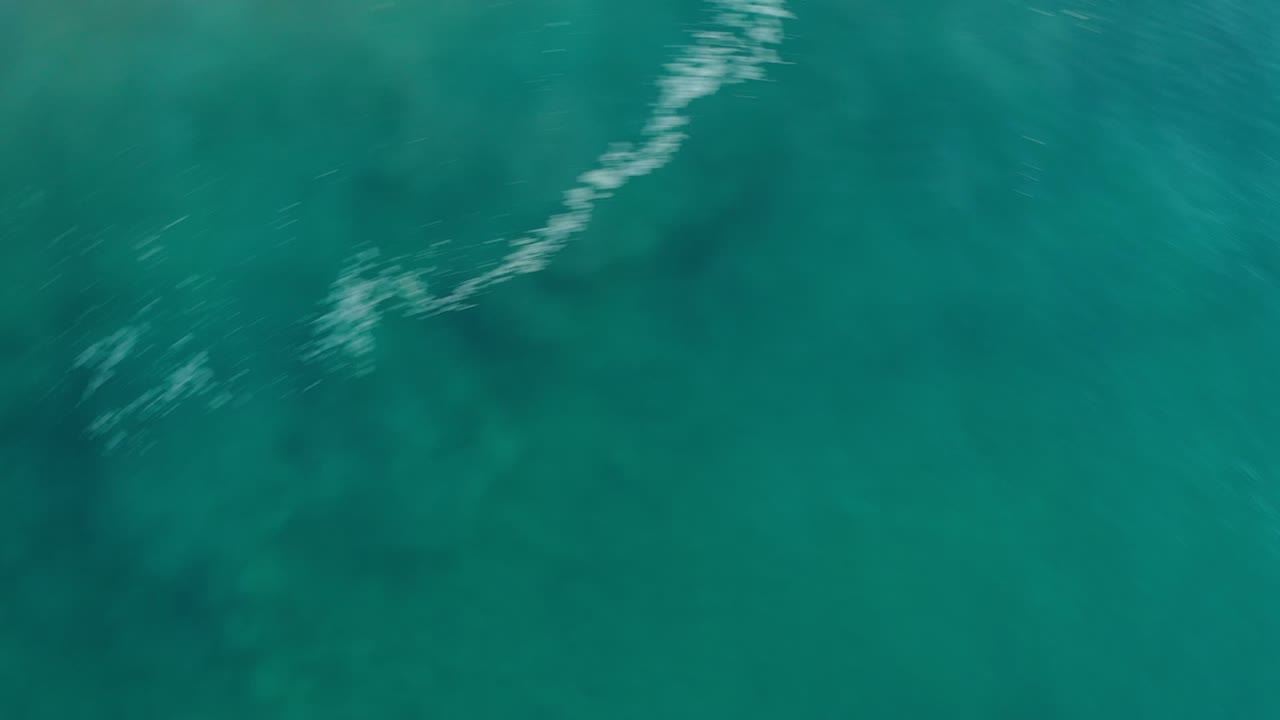 Clear Los Cabos ocean water from above, with reef shadows under the surface and a pale white wave line entering near the top edge, no visible people.