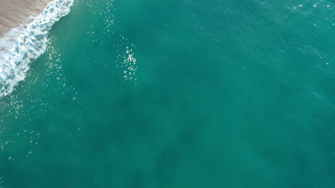 Aerial shoreline frame from Los Cabos, Mexico, showing white foam crossing tan beach beside clear turquoise water with no visible people.
