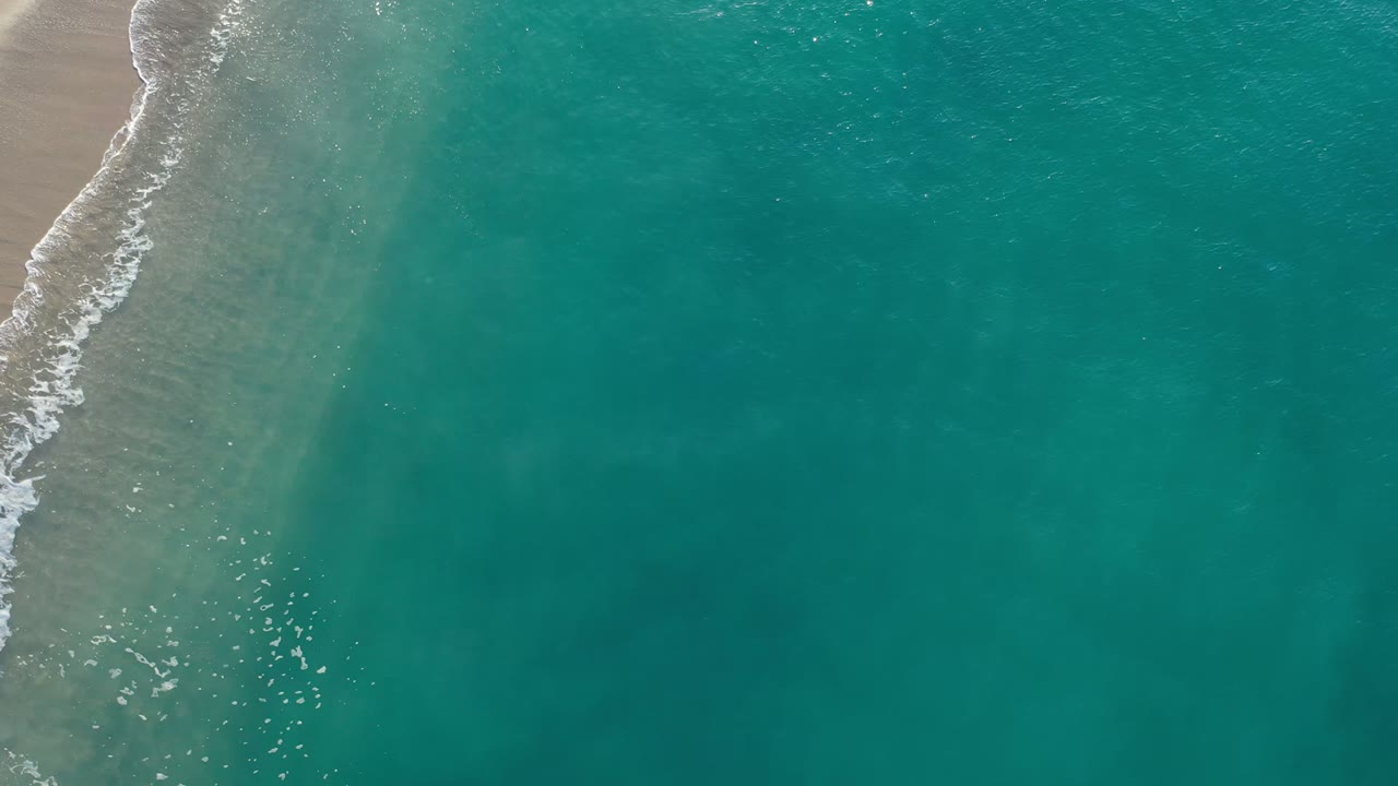 Aerial view of teal ocean beside a narrow sandy shoreline.
