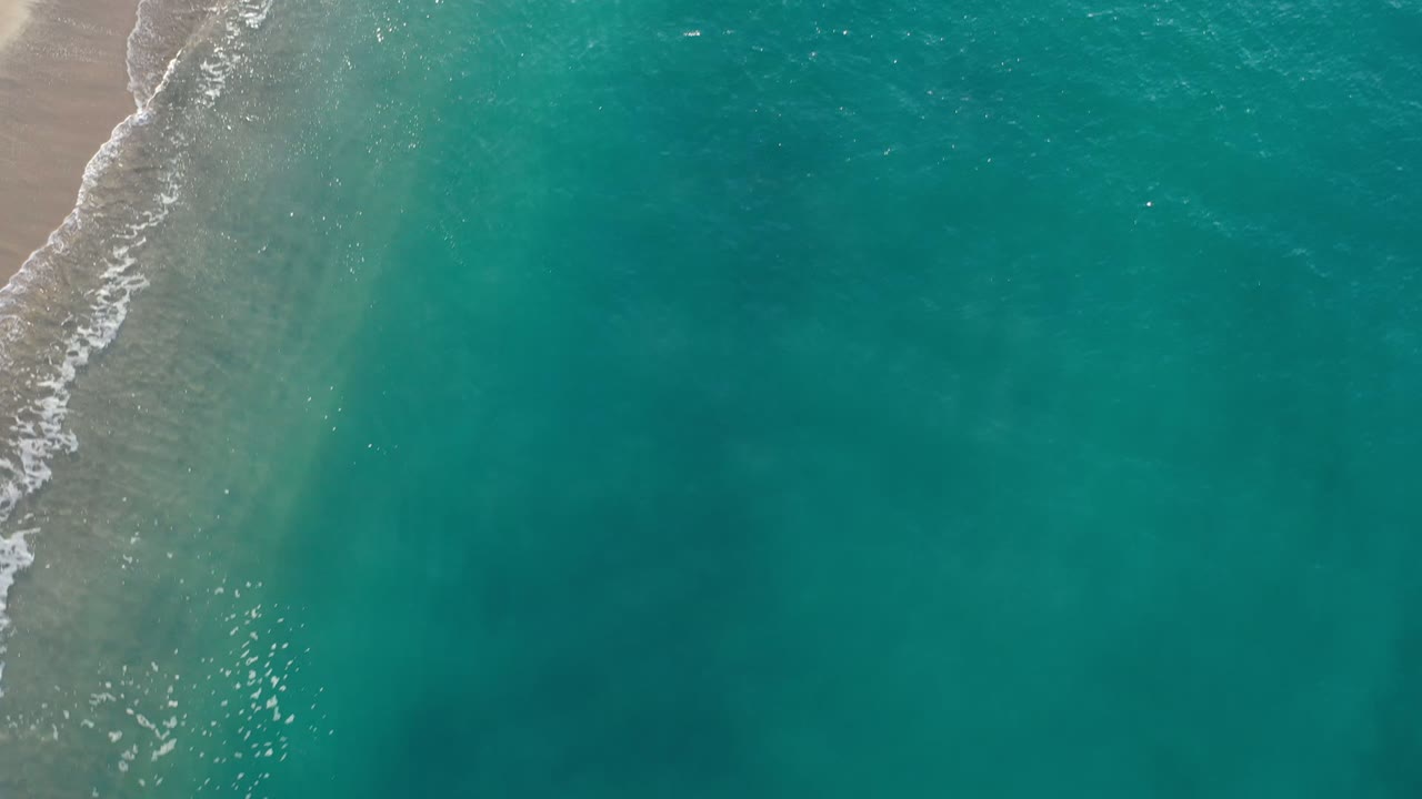 Overhead Los Cabos beach frame with the surf line moving down the left edge and clear teal water spread across most of the image, no visible people.