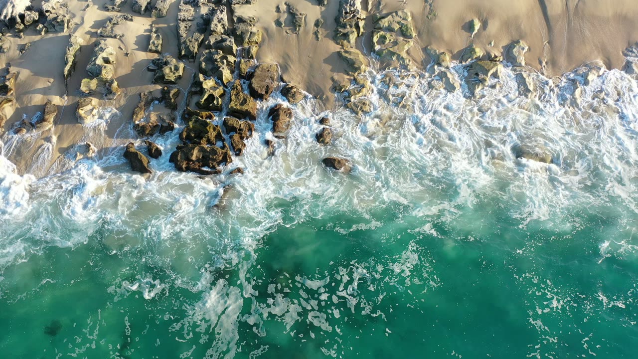 Overhead surf breaking on rocks and sand beside turquoise water.