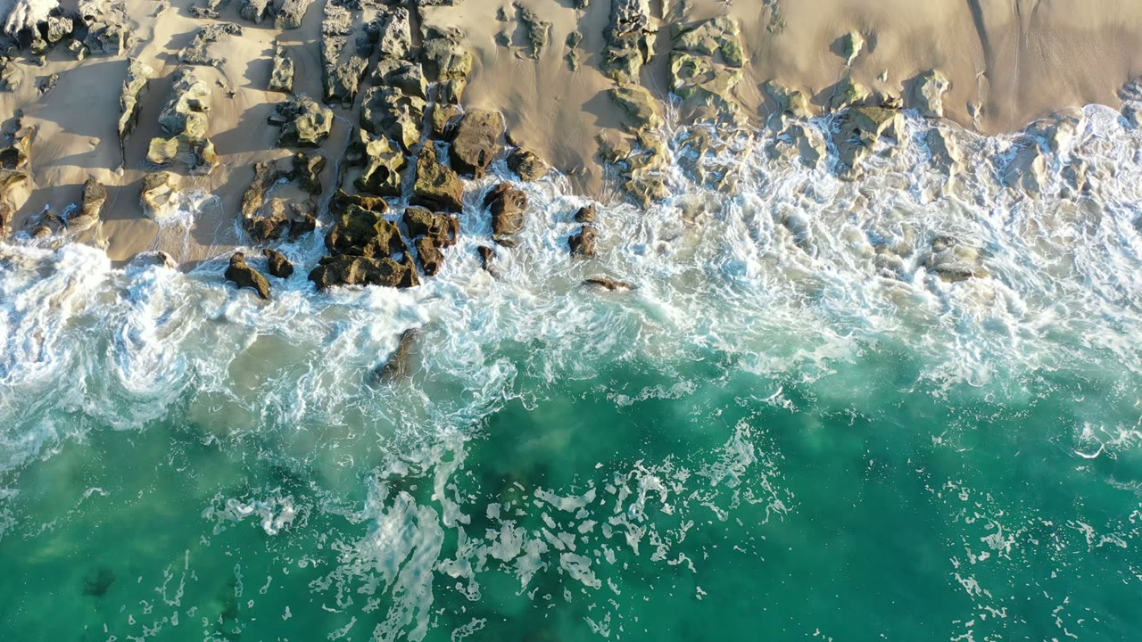 Overhead Los Cabos beach frame with a broader band of white foam washing around exposed rocks and green-blue water in the foreground, no visible people.