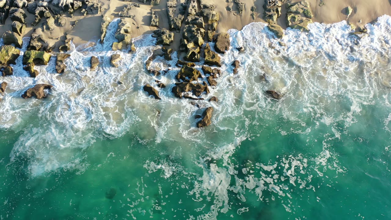 Overhead view in Los Cabos, Mexico, with green ocean water below and white foam pushing into dark shoreline rocks and pale sand, no visible people.