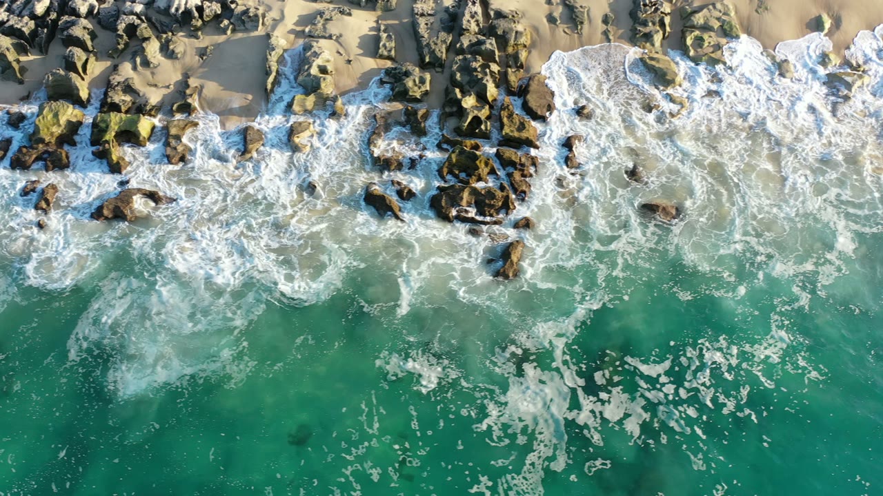 Aerial frame in Los Cabos, Mexico, showing white surf spreading over scattered dark rocks with green water in the lower half and sand along the top, no visible people.