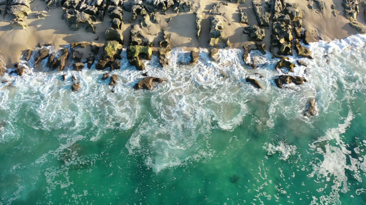 Overhead surf breaking over rocks beside a sandy beach.