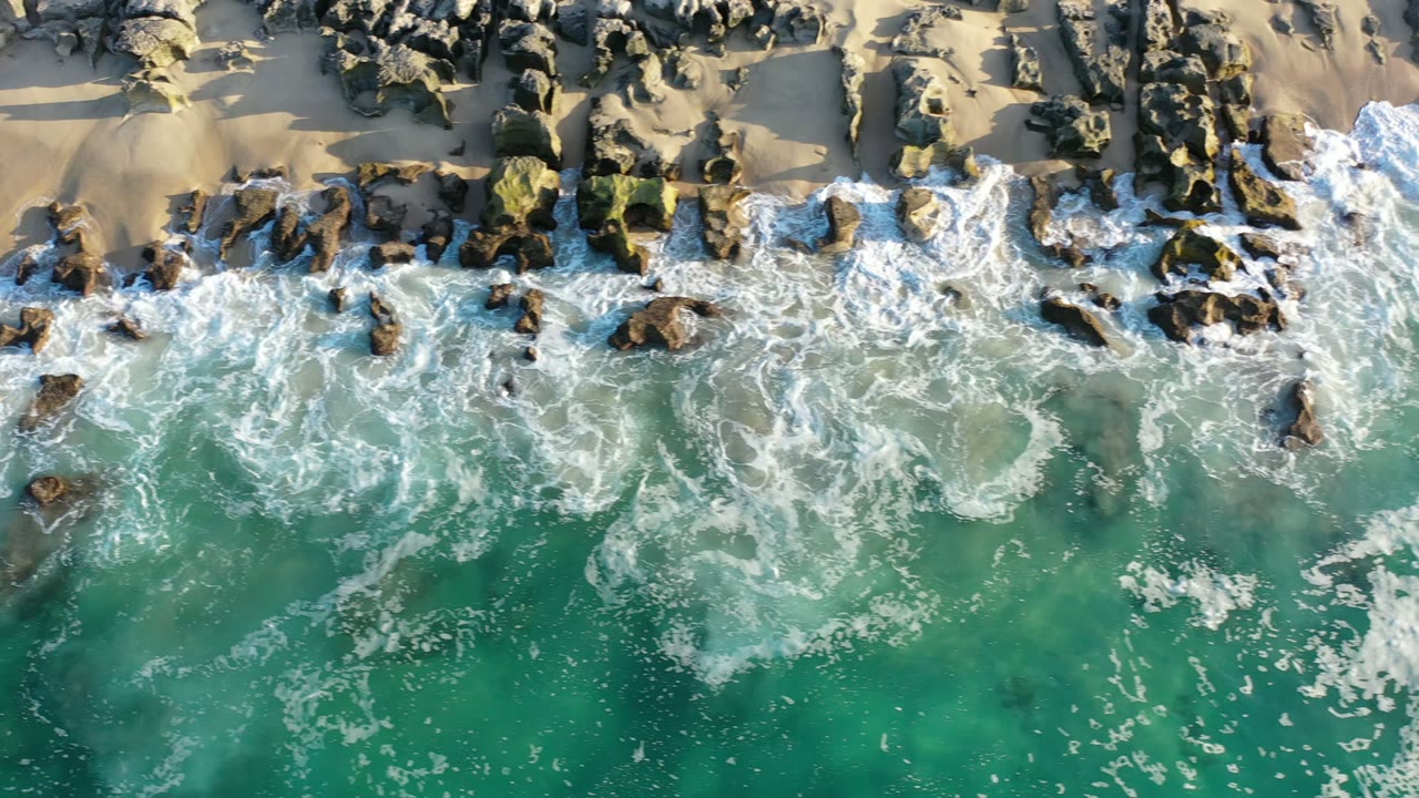 Los Cabos aerial shoreline frame with foamy waves spreading across scattered rocks and clear green-blue water, with no visible people in this frame.