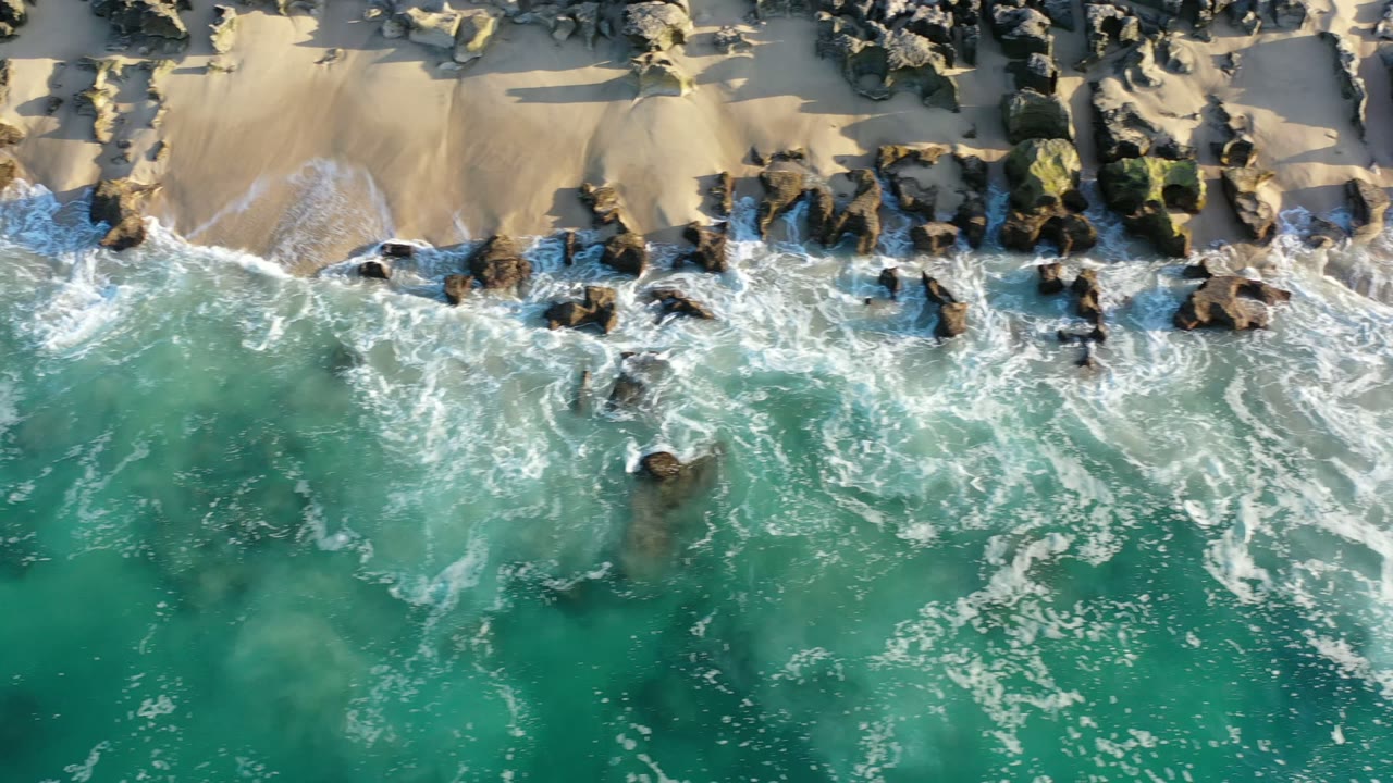 Overhead view in Los Cabos, Mexico of turquoise water pushing white foam toward dark shoreline rocks and pale sand, with no visible people.