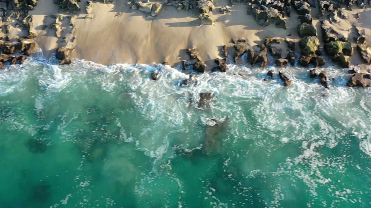 Aerial Los Cabos shoreline frame with whitewater spreading across scattered rocks while clear green-blue water fills the lower half and no visible people appear.