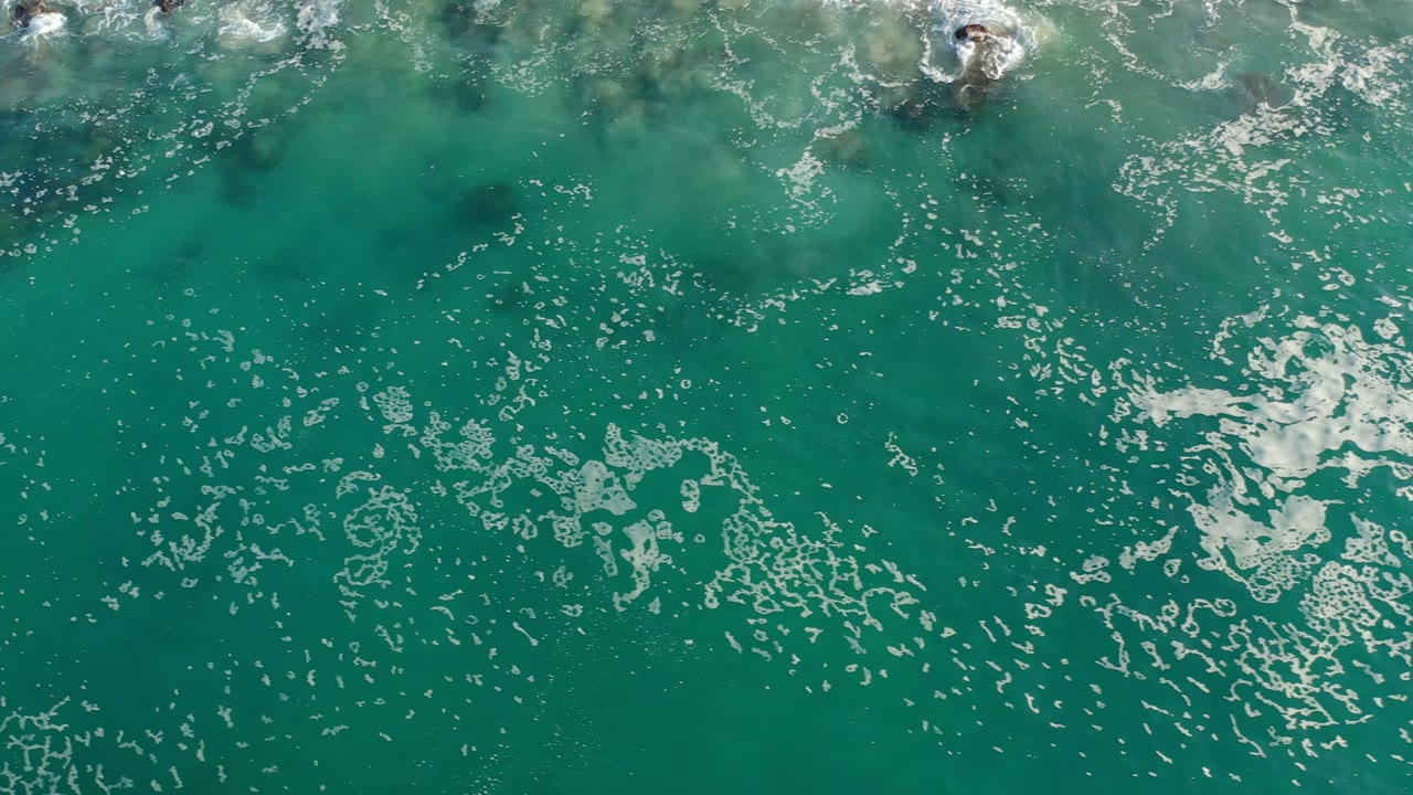 Overhead Los Cabos ocean frame with a bright white foam patch near center, teal water around it, faint darker areas below the surface, and no visible people.
