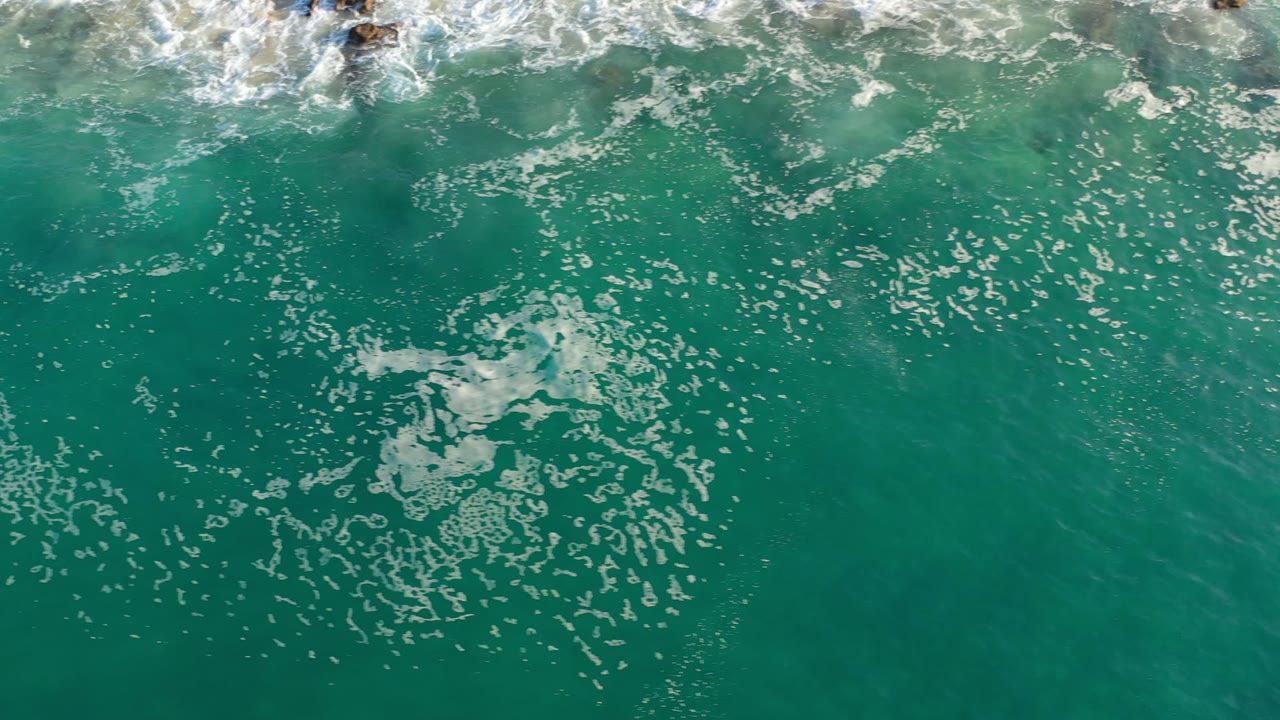 Overhead view of green seawater in Los Cabos with white bubbles spread across the surface, breaking foam along the top edge, and no visible people.
