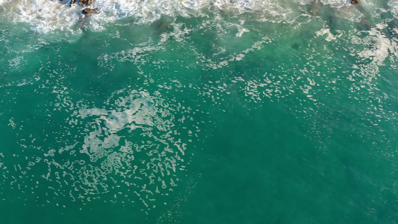Aerial view of Los Cabos green ocean water with a loose cluster of white bubbles near center frame and foamy surf touching the upper edge, no visible people.