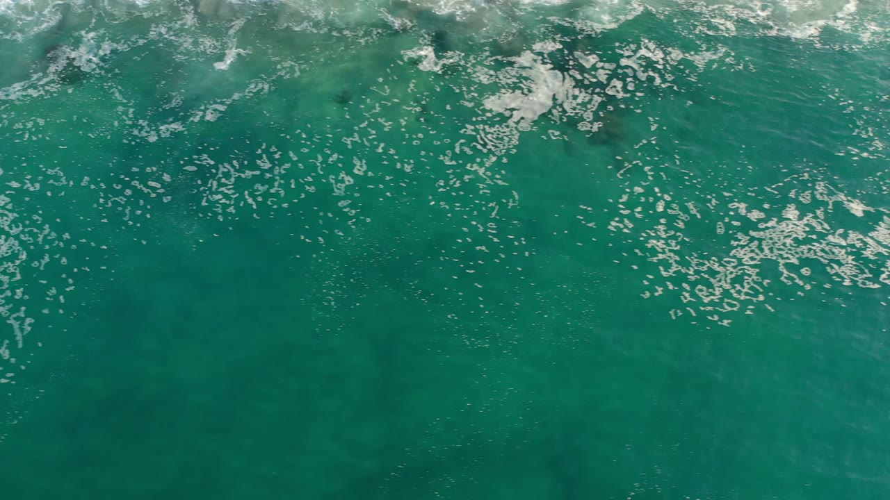 Overhead view of turquoise water and scattered white foam over submerged rocks in Los Cabos, Mexico, with no visible people.