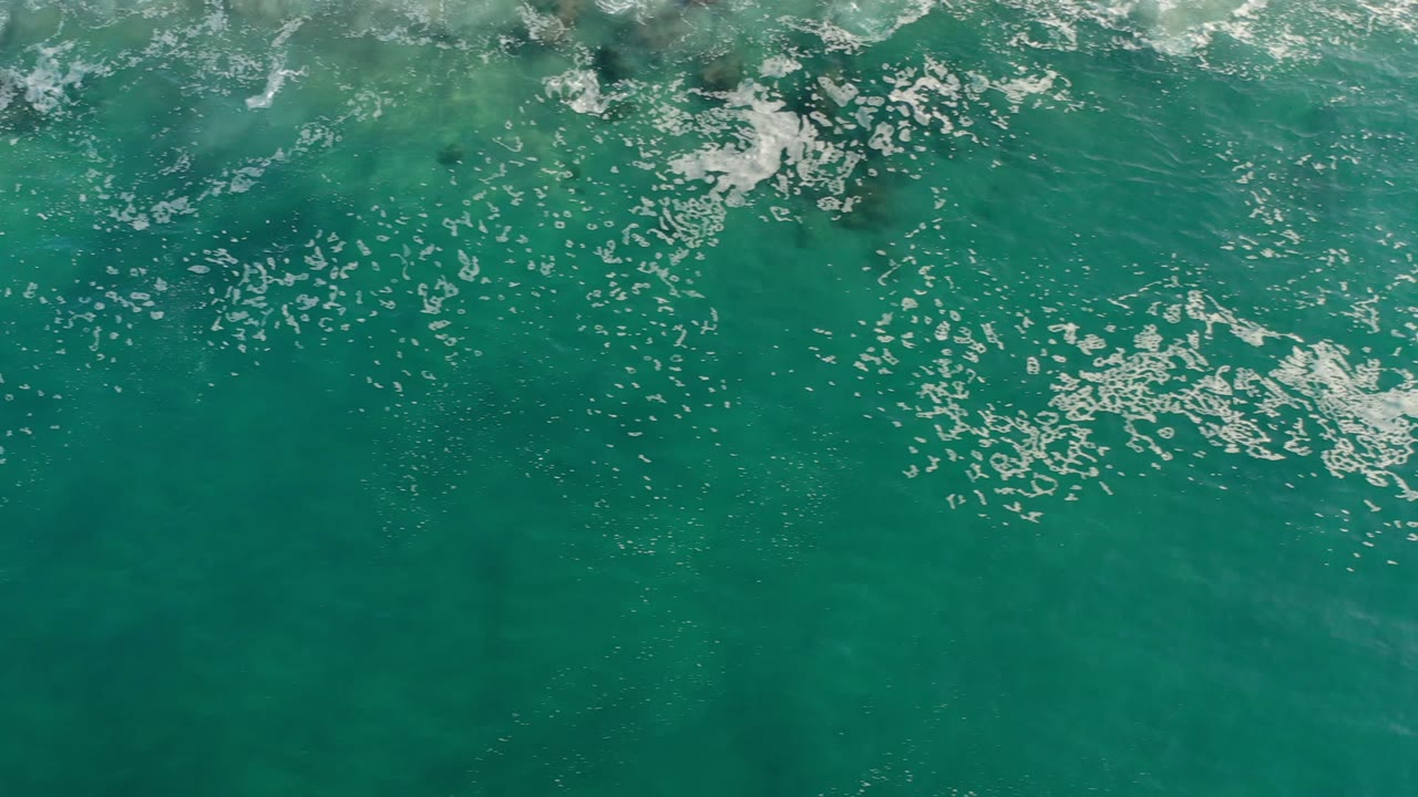 White foam spreads across green-turquoise water near darker rock shapes in Los Cabos, Mexico, with no visible people.