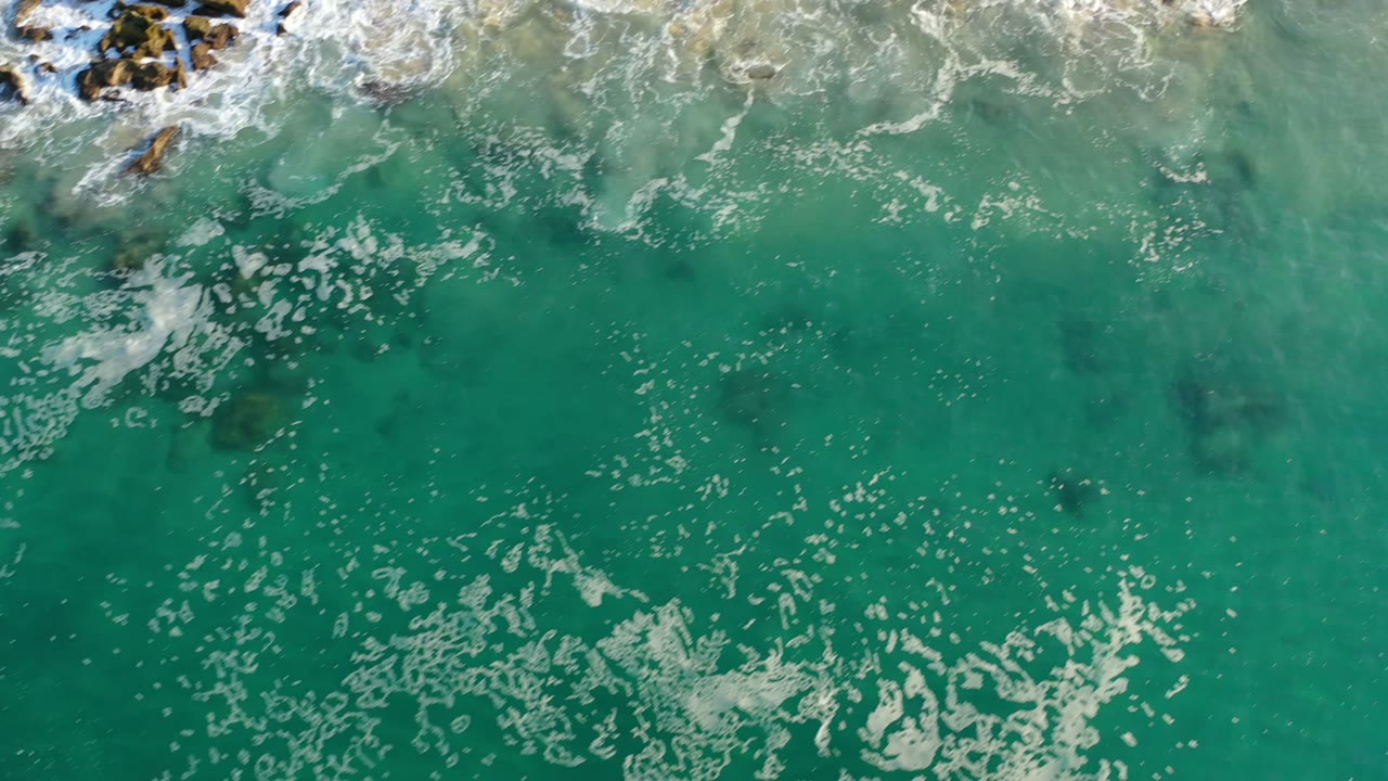 Aerial Los Cabos frame looking down through clear green water, with scattered foam streaks and a cluster of rocks breaking the surface near the top, with no visible people.