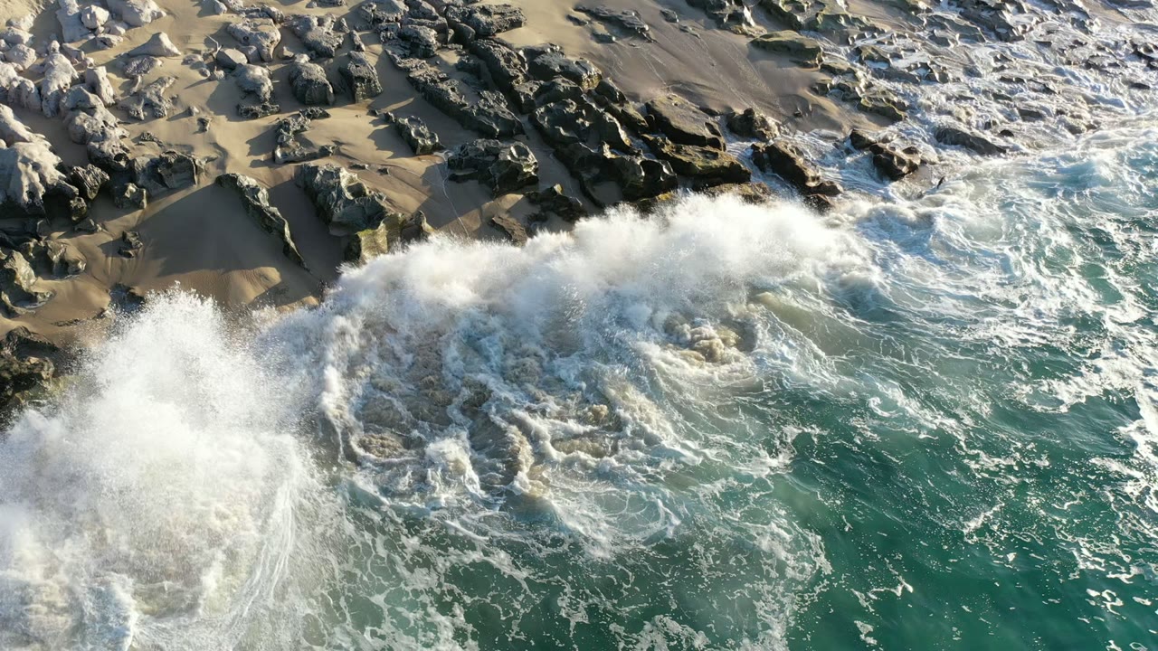 Aerial view in Los Cabos, Mexico of white surf striking dark rocks beside green water and tan sand, with no visible people.