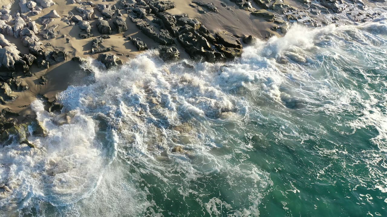 Aerial Los Cabos shoreline view with white foam spreading over dark rocks and green sea water, with no visible people.