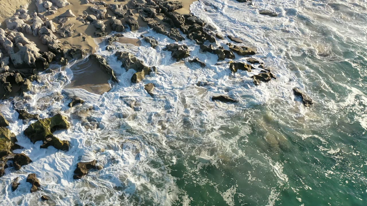 Overhead view of Los Cabos surf spreading across dark shoreline rocks and wet tan sand, with green-blue water below and no visible people.