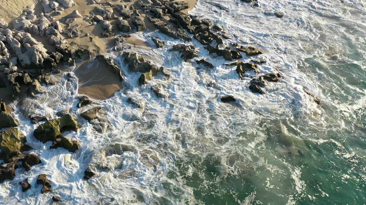 Los Cabos shoreline seen from above as white foam threads between dark rocks, with a small wet sand pocket near the upper center and no visible people.