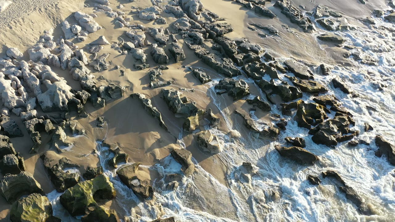 Foamy ocean water spreads across uneven dark rock shelves and pale wet sand in Los Cabos, Mexico, with no visible people.