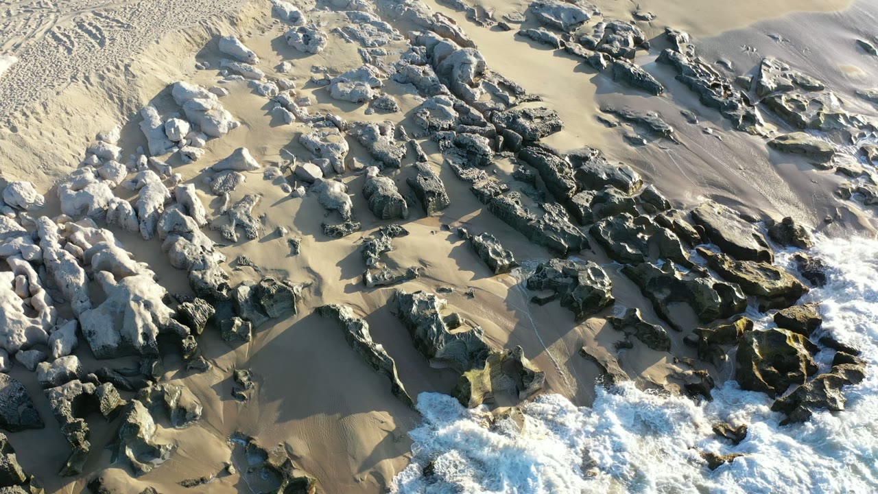 Overhead view of a rocky beach in Los Cabos, Mexico, with tan sand, dark rock formations, white surf at the lower edge, and no visible people.