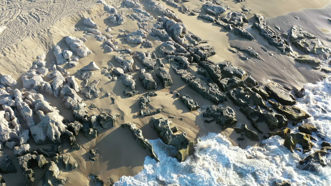 Top-down frame of Los Cabos beach rocks set in wet and dry sand, with white water entering from the lower right and no visible people.