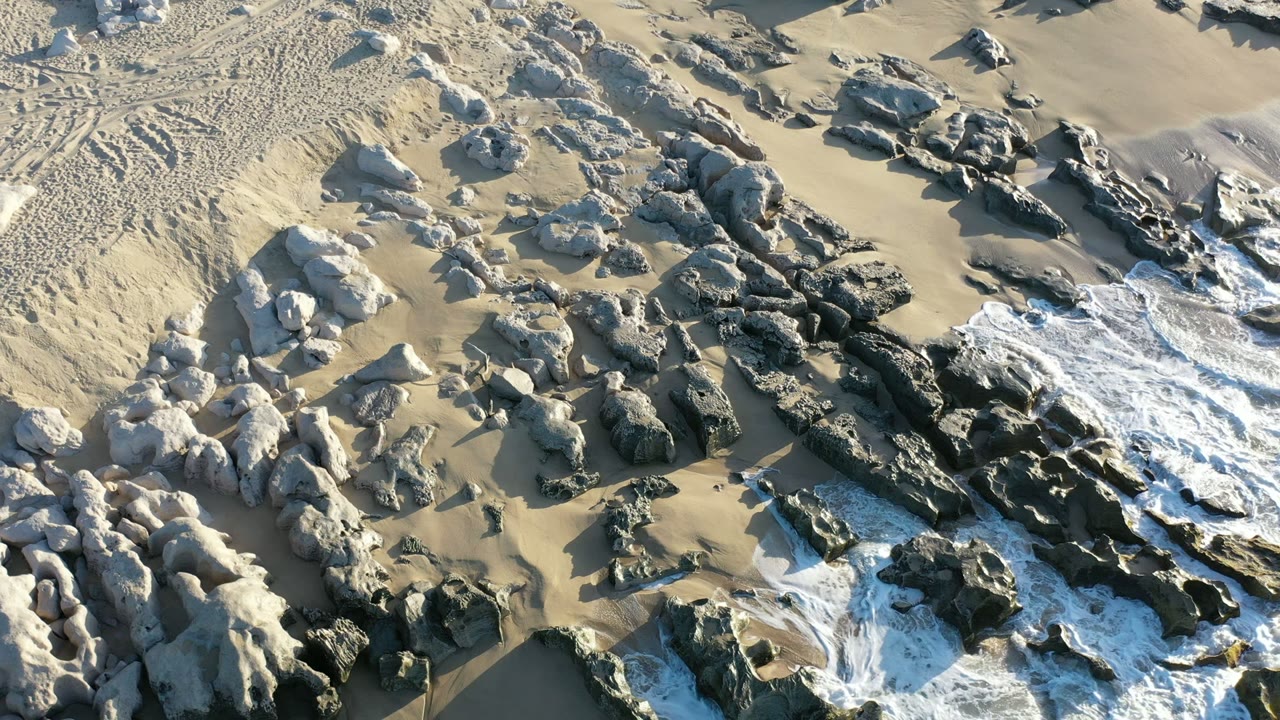 Overhead view of rocks, pale sand, and sea foam along a beach.