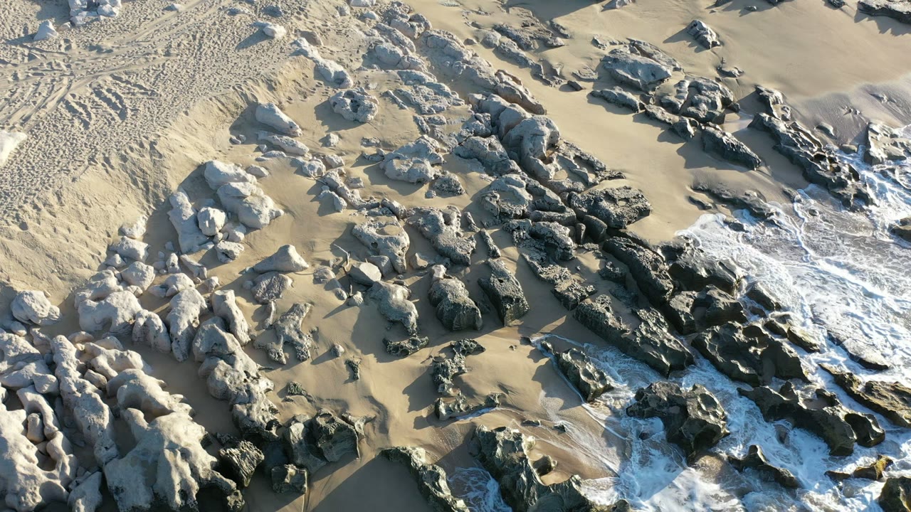 Overhead Los Cabos shoreline view with darker central rocks, thin foam trails, wet sand near the lower edge, and no visible people.