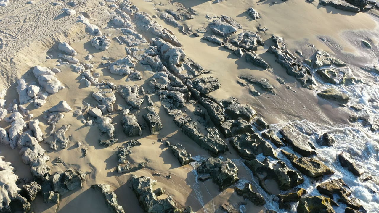 Overhead view of a Los Cabos rocky shoreline with pale sand, dark rocks, white foam at the lower right, and no visible people.