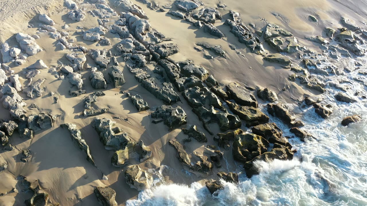 Overhead Los Cabos beach view with white surf entering from the lower edge, dark rock clusters across tan sand, and no visible people.