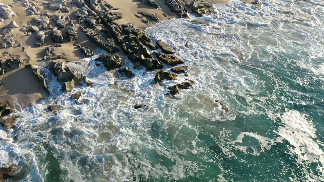 Aerial frame from Los Cabos, Mexico showing foamy white waves spreading across dark shoreline rocks, turquoise water in the lower frame, tan sand above, and no visible people.