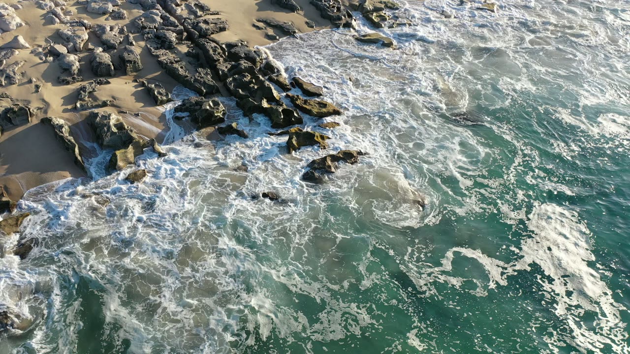 Aerial Los Cabos shoreline frame with whitewater curling around scattered dark rocks, sandy gaps along the top edge, teal water below, and no visible people.